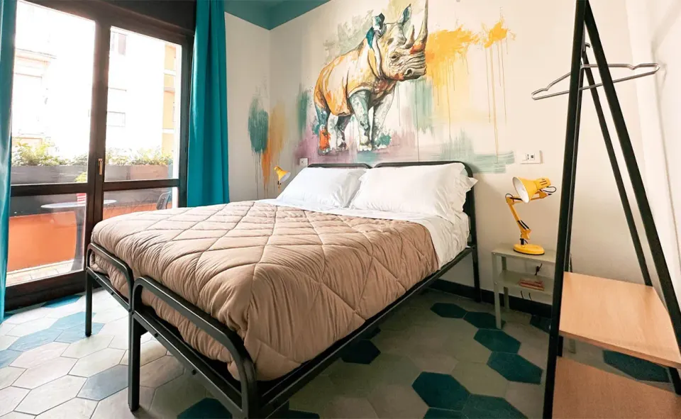 A bedroom with a double bed, teal tiled floor and a painted rhinoceros mural on the wall beside a large window