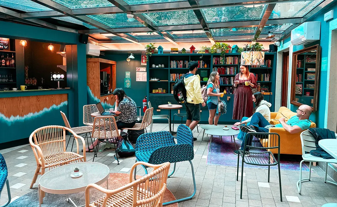 The glass-roofed lounge and bar of the hostel, with rattan chairs, bookshelves and guests chatting at small tables